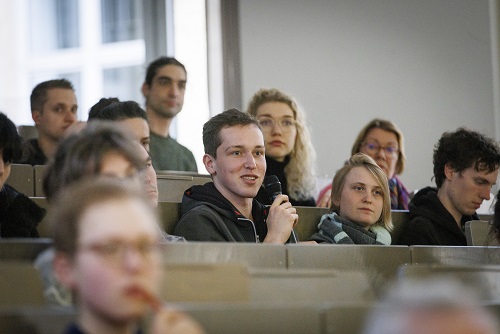 Student stellt Frage während Vorlesung in Hörsaal mit mehreren Zuhörern.