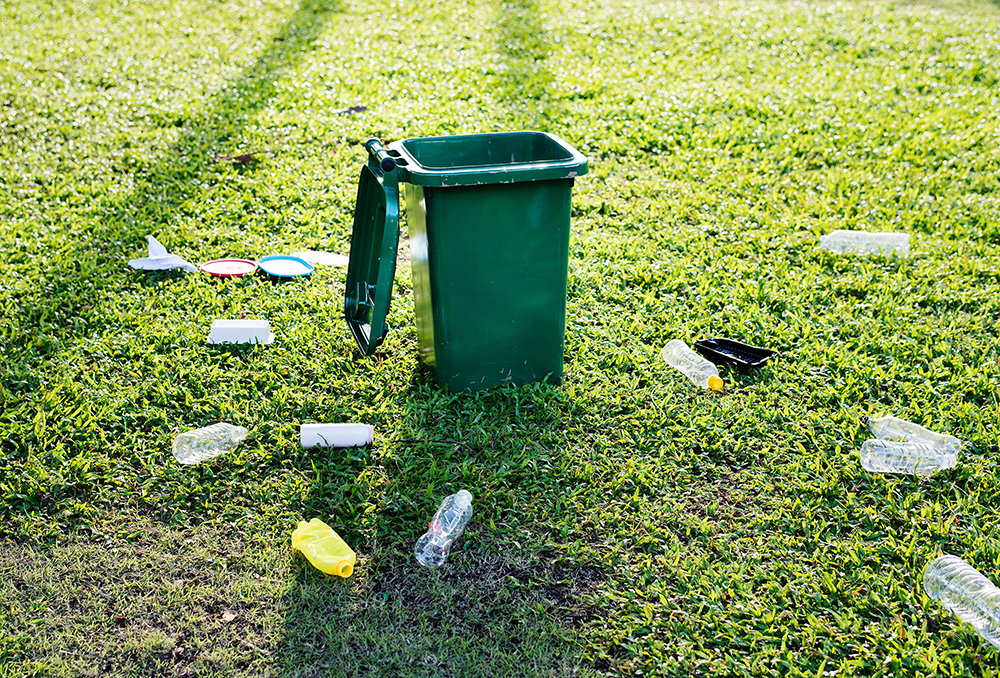 Offene Mülltonne auf Wiese mit Plastikmüll darum verstreut